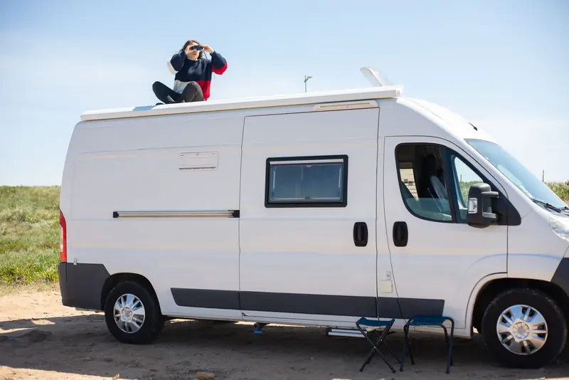 A woman sitting on the roof of a camper van using binoculars during a sunny day