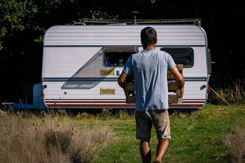 A man carrying a wooden crate approaches a caravan in a sunlit grassy area, evoking a rustic outdoor lifestyle